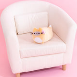 Pusheen Enchanted Fox plush sits on a cream chair to demonstrate the plush’s size. The orange, white, grey, and pink plush has brown embroidery features for Pusheen's eyes, mouth, and whiskers. 