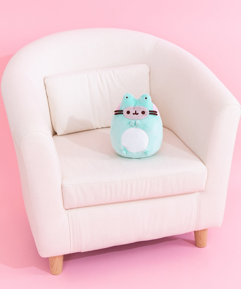 Pusheen Enchanted Frog plush sits on a cream chair to demonstrate the plush’s size. The green, white, grey, and pink plush has brown embroidery features for Pusheen's eyes, mouth, and whiskers. 