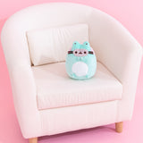 Pusheen Enchanted Frog plush sits on a cream chair to demonstrate the plush’s size. The green, white, grey, and pink plush has brown embroidery features for Pusheen's eyes, mouth, and whiskers. 