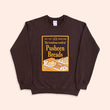 A chocolate brown sweatshirt with the sleeves folded lies against a light-yellow background. The unisex sweatshirt has a screen-printed graphic on the center of the chest. The graphic depicts a magazine cover showcasing “the wondrous world of Pusheen Breads.” This is a special, first edition cover featuring Loaf Pusheen, Baguette Pusheen, Croissant Pusheen, Loaf Pusheen, and Sesame Bagel Pusheen in golden shades of baked goodness. 
