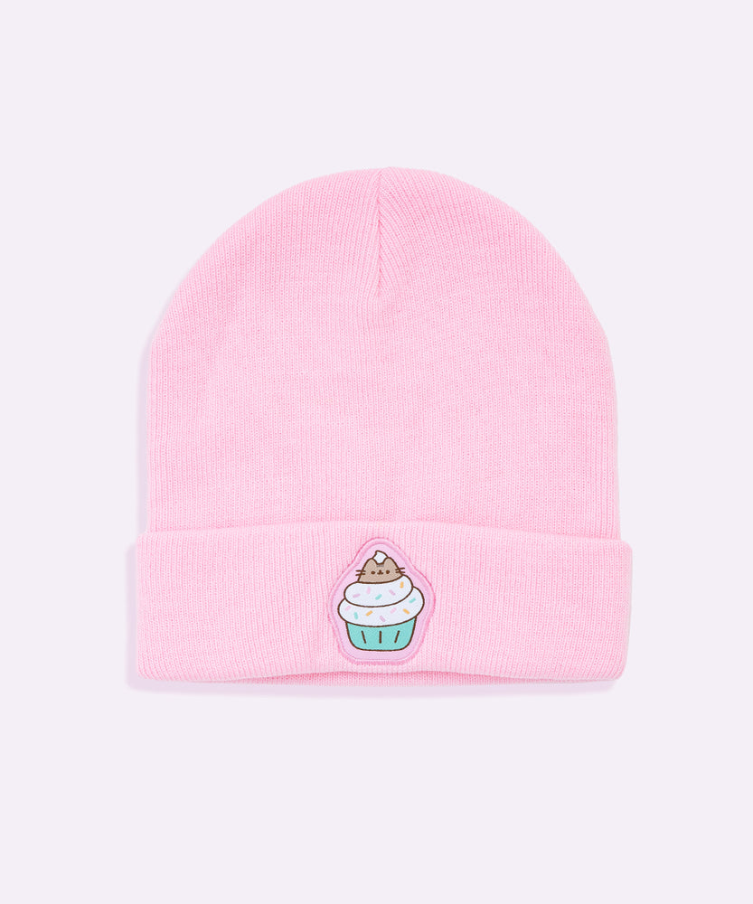 Front view of the Pusheen Cupcake Knit Hat. The light pink beanie lies on a white surface. On the front center of the folded edge is an embroidered Pusheen the Cat sitting inside the whipped icing of a cupcake. With multi-colored sprinkles and a teal base, the patch is surrounded by a light pink background. 