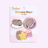Pusheen Dreamy Days Pin Set lies in front of a light purple backdrop. Three Pusheen pins are attached to a backer card with the product name in gold foil print. The top left pin shows Pusheen resting on a fluffy white cloud. The top right pin shows Pusheen lying in a circle while the phrase “Lazy Club” is to the side of Pusheen in gold foil. The bottom pin shows Pusheen holding a shooting star with light pink, pink, and light purple streaks coming off the yellow star. 
