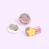 Close-up view of the Pusheen pins on a light purple background. The gold foil outline of the pins is hitting the light to show its reflective property.  