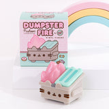 Front view of Pusheen Dumpster Fire Vinyl Figure. The toy is placed in front of its packaging box, which features a design of the trash toy surrounded by a donut, candy, and chocolate. The front of the packaging reads “Dumpster Fire by 100% Soft, Pusheen Flocked Edition, Vinyl Figure.” The main colors included on the packaging are light pink, mint green, grey, and light brown. 