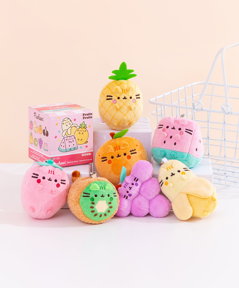 Front view of Pusheen Fruits Surprise Plush assortment. Plush keychains sit on multi-level white pedestal in front of a light-yellow background. Keychains feature various fruits including a pineapple, strawberry, orange, kiwi, grapes cluster, watermelon slice, and peeled banana. Accompanying the seven fruits keychains is the mystery box packaging for the Fruits Surprise Plush. 