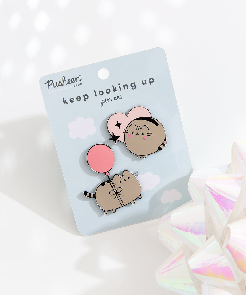 Two Pusheen pins are attached to a light blue backing card with the Pusheen Shop logo and the pin set name, “keep looking up pin set,” in a dark blue color. The pin set lies on a white background with a reflective white bow in the right corner. 