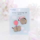 Pusheen Keep Looking Up Pin Set lies in front of a white backdrop with reflective decorative bows. The Two Pusheen pins are attached to a backer card with the product name in dark blue color. The top right pin shows a round blushing Pusheen with a pink heart and two sparkles. The bottom left pin shows Pusheen attached to a pink balloon and floating.  