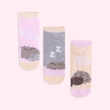 Pusheen Nap Club Socks lie on a light pink surface. The sock set features the medium grey Pusheen the Cat with her siblings: a small gray cat named Stormy and a dark brown cat named Pip. 