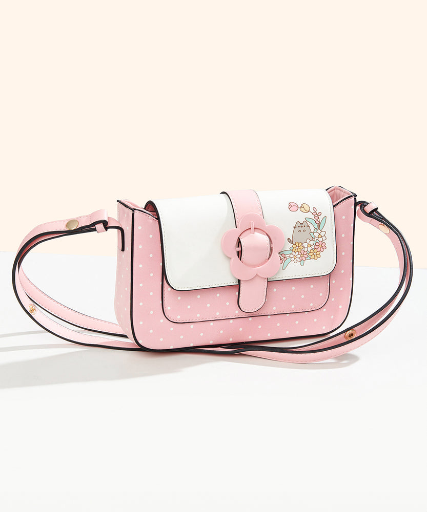 Front view of the Pusheen Springtime Purse. The pink rectangular shaped purse has rounded edges. Attached to the pink base with white polka dots is a light pink strap that matches the purse color. On the closure flap is a pink daisy buckle with a white portion. On the white portion is a graphic of Pusheen with flowers.