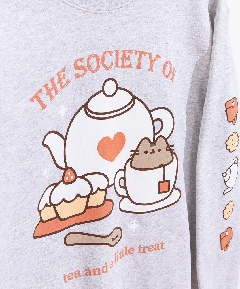 Close-up view of the front and sleeve graphics of the grey sweatshirt. Pusheen the Cat sits in a white teacup surrounded by a white tea kettle and cupcakes. The phrase “The Society of tea and a little treat” is printed above and below the Pusheen graphic in a salmon color. 