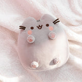 Toe beans plush lays on a white fluffy surface while the sun shines on half of the plush, highlighting the light grey coloring of the plush. Pusheen’s brown whiskers extended off the front side of the plush. 