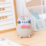 Front view of Gaming Pusheen Lamp. The grey tabby cat is holding a blue, pink, and orange gaming device in her paws while a pair of pink and blue headphones sit atop her ears. The cat’s facial features are navy blue while her head stripes are dark grey.  