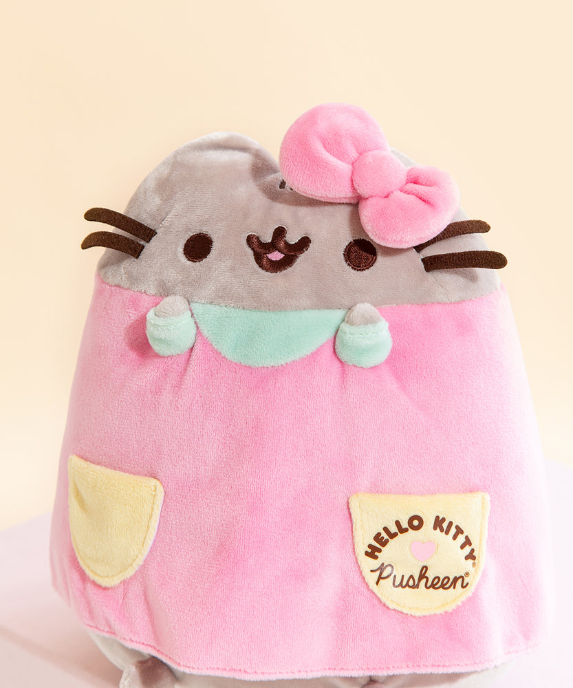 Closeup of the front of the plush. The Hello Kitty x Pusheen collaboration logo is screen printed onto the dress pocket, while Pusheen's face is embroidered on. The bow on Pusheen's right ear covers up her ear.