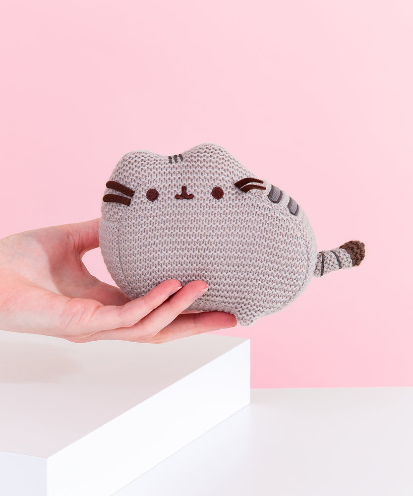 Model holds Knit Pusheen Plush. Mini plush is made with a grey knit fabric thread to give the plush a crocheted look. Grey embroidery is used for Pusheen’s stripes while brown embroidery is used for Pusheen the Cat’s mouth and eyes. Plush measures 6” long and 4” tall.  