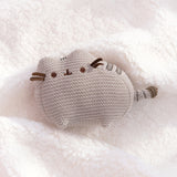 Knit Pusheen Plush lies on a white, fluffy surface. The brown ribbon whiskers extend off the plush face. The plush is sculpted so the cat’s ears and paws are defined. 