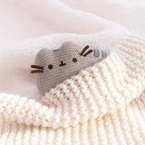 Mini Plush lies on a white, fluffy surface with a cream knit blanket covering half of the plush. The crocheted plush is snuggled into the blanket. 