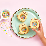 Top view of a trio of Pusheen cookies with a see-through stomach with sprinkles inside. A model’s hand holds one of the cookies above the mint and gold plate, and a scoop of sprinkles has slightly spilled to the left.