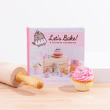 A square hardcover book standing on top of a wooden board, surrounded by a rolling pin, a pink cupcake, and a white pedestal. The book reads ‘Let’s Bake! A Pusheen Cookbook’ and features a photo displaying all the Pusheen treats you can bake with the book. The author names Claire Belton and Susanne Ng are noted in pink at the bottom.