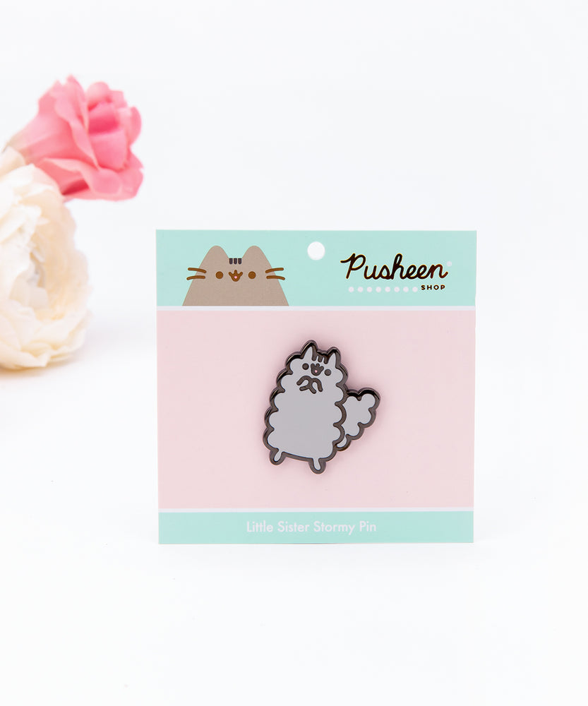 The Stormy pin in it’s packaging, laying on a white floor, a pair of pink and white peonies partially visible in the top left corner. The packaging is a square cardboard backing, a pink square in the middle and mint banners on the top and bottom. The top banner features the Pusheen Shop logo, and the bottom banner says ‘Little Sister Stormy Pin’ in white.