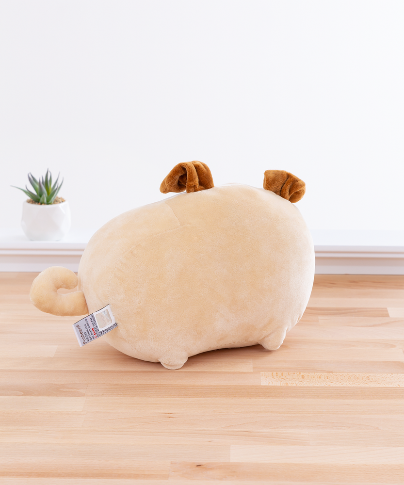 Back view of the Pusheen Plush in front of a white wall on top of a wooden floor, with a potted succulent to the plush’s left. There are no details on the plush’s back, and the information tag on the plush is nearby the curly tail. The back view makes it more obvious Pugsheen’s ears are folded over.