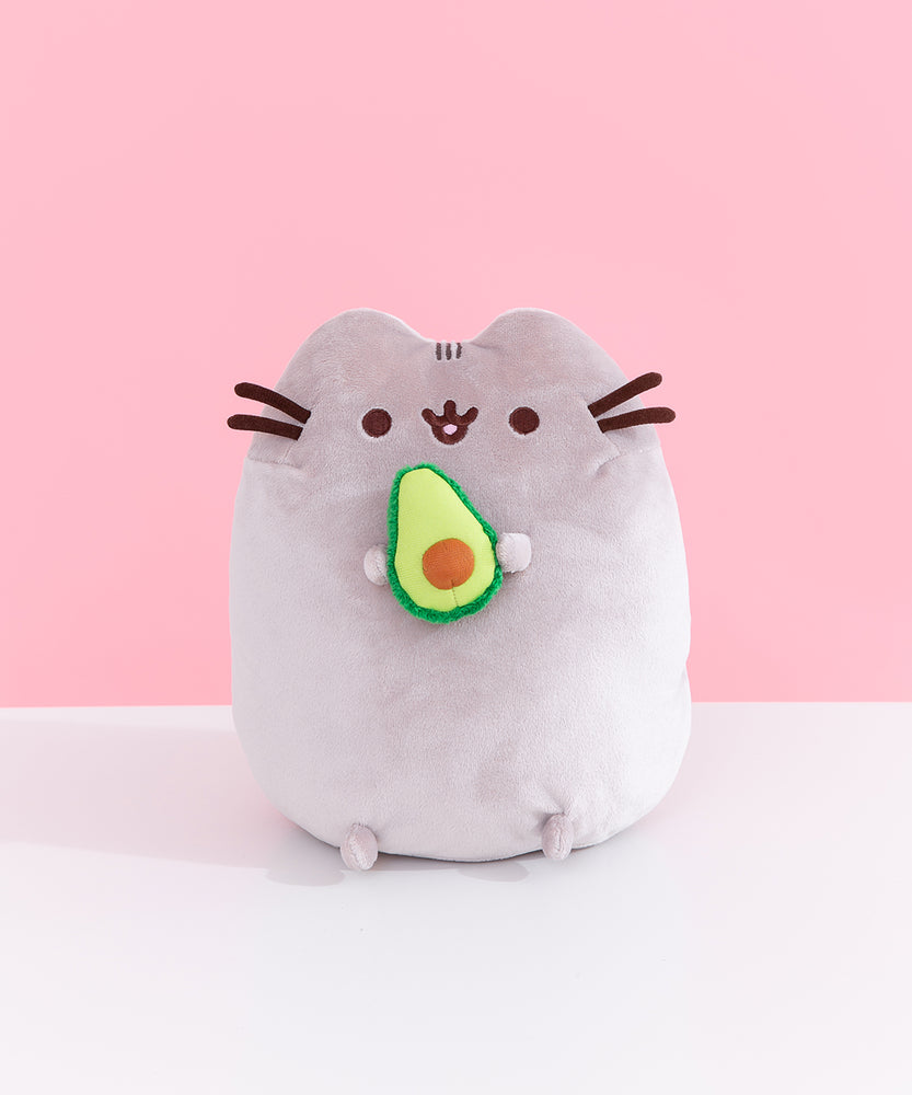 Front view of Pusheen Avocado Plush. Pusheen the Cat is shown with her classic brown eyes, mouth, and whiskers. Pusheen holds a cut-in-half, two-toned green avocado with a light brown pit.