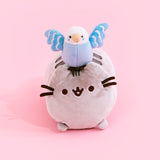 Plush of Bo sitting on Pusheen's head. Bo is a light blue parakeet with a white head, yellow beak, dot eyes and blue dots on her cheek. Bo's darker blue wings are spread out wide with three rows of white feathers, and her dark brown feet are perched in between Pusheens' ears. Pusheen is shaped like a log, laying on her stomach and her paws laying out from the bottom.