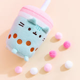 Close-up view of the Boba Tea Sips Plush. On a light-yellow surface, the plush is laid down on its back with its feet up and is surrounded by multi-color pink pom-poms. The bottom of the plush is flat, like an actual cup. 