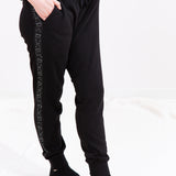 Model wearing slightly baggy black joggers. The side of the jogger features a strip of black fabric with a pattern alternating between white star constellations and white line art of Pusheen hopping. The model has a hand in the pocket of the jogger.