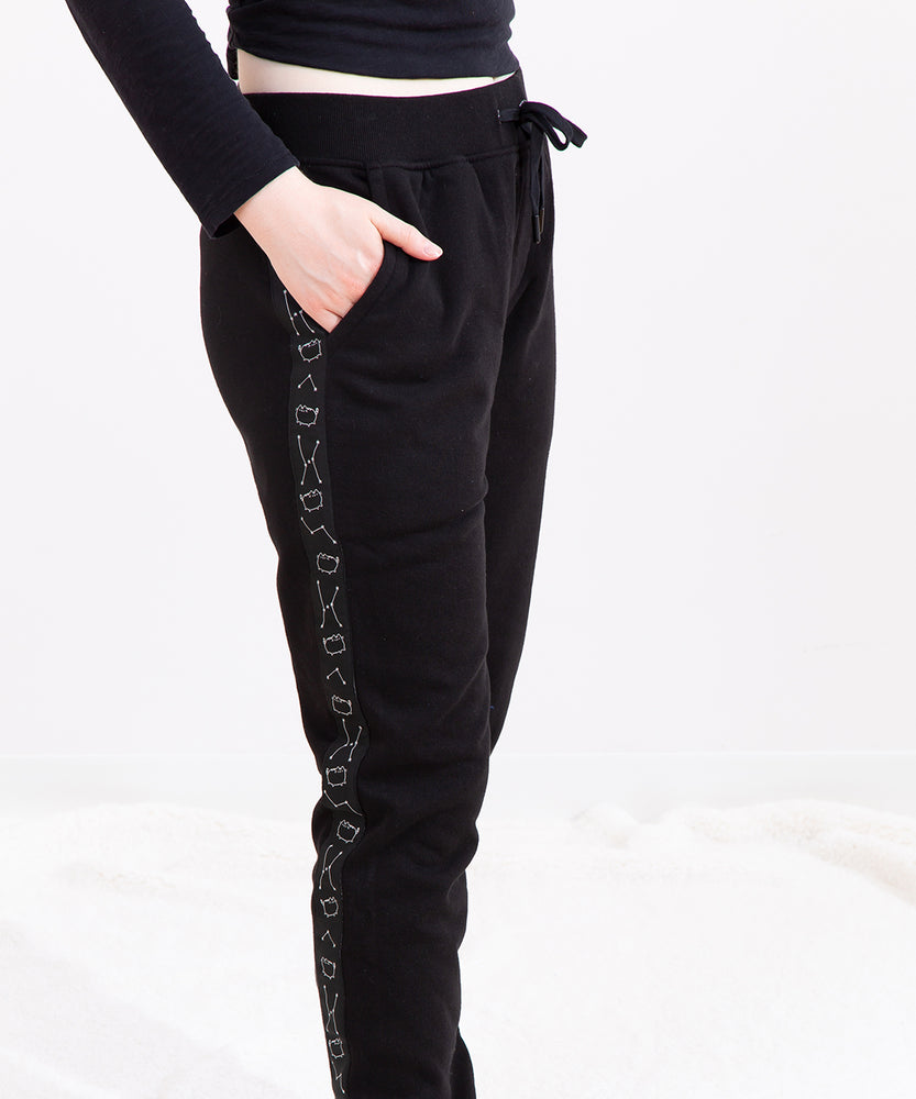 Model wearing the Pusheen Celestial Unisex Jogger, showing the pattern on the side of the jogger with a hand in the jogger's pocket. The jogger has an elastic waistband with a black drawstring tied into a bow. The model is standing in front of a white background.