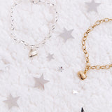 Gold and Silver charm braclets next to one another on top of a plush white blanket and surrounded by silver stars. The charm features Pusheen sitting in a loaf pose, with her feet tucked in and her tail by her side. The loops on the chain link are larger then normal.