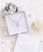 The Silver Charm Bracelet in a white square jewelry box. The box is being propped up by a shiny silver gift bow, and the scene is decorated with silver stars, the lid of the jewelry box, and a white holiday ornament, all on top of a plush white blanket.