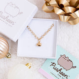 The Gold Charm Bracelet in a white square jewelry box. The jewelry lid is partially on top of the box, and the Pusheen the Cat cart insert for the jewelry box is underneath the bottom right of the box.The box is surrounded by a shiny gold gift bow and white and bronze holiday ornaments, all on top of a plush white blanket.