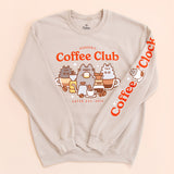   Front and sleeve view of the tan unisex sweatshirt. The arms are folded in to the center of the sweatshirt to reveal the printed sleeve detail. The sleeve features the phrase “Coffee O’ Clock” with Pusheen sitting in a white coffee cup surrounded by two coffee beans.   
