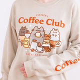 Model wearing the Pusheen Coffee Club Unisex Sweatshirt. The tan sweatshirt features a large graphic in the center of the sweatshirt of Pusheen the Cat, Little Brother Pip, and Little Stormy enjoying coffees and sweet treats. The phrase “Pusheen’s Coffee Club” sits above the cats in red orange text while the phrase “Catfé Est. 2010” sits below the graphic. The model is holding their arm up to show a printed sleeve detail featuring the phrase “Coffee O’Clock”. 