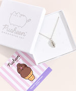 Sterling silver finish of Pusheen Dipped Cone Charm Necklace in its white packaging box. The necklace is hanging inside the white box with a pink, white, and purple informational card. 