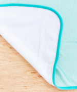 Close-up view of reverse side of blanket. The teal velour blanket is backed with a white velour. 
