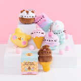 Stylized front view of Pusheen Ice Cream Surprise Plush assortment sitting on multi-level white pedestal in front of a pink background. Accompanying the seven characters of ice cream Pusheens is the mystery box packaging for the Pusheen Ice Cream Surprise Plush. 