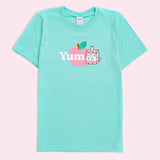 A mint green graphic tee against a light pink background. The graphic features Pusheen as Juice Box Sips next to the word ‘Yum’ in white, both in front of a pink apple. 
