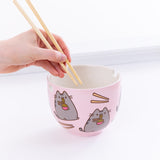 A hand holding the chopsticks over the Pusheen Ramen Bowl from the left, in front of a white background. 