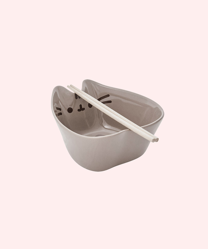 Right quarter view of the Pusheen Ramen Bowl & Chopstick Set. The grey bowl is in the shape of Pusheen the Cat and features her signature whiskers, mouth, eyes, and stripes printed on the interior of the bowl. The included chopsticks rest in the divot between Pusheen's structured ears. 