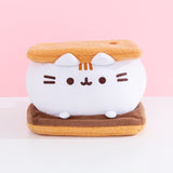Front view of Pusheen S’mores Squisheen. Pusheen the Cat takes the form of a delicious campfire snack: a s’more. Pusheen's body is the white middle marshmallow. The white marshmallow sits on a dark brown slab of chocolate. The marshmallow is sandwiched between two light brown graham crackers inspired plush.