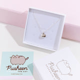 The silver charm necklace in a square white jewelry box, propped up against a pink pedestal, the square card insert and the lid of the box laying underneath it. The square card features an illustration of the Pusheen the Cat logo.