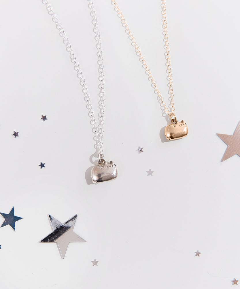 Gold and Silver charm necklaces next to one another on top of a white blanket and surrounded by silver stars. The charm features Pusheen sitting in a loaf pose, with her feet tucked in and her tail by her side. 