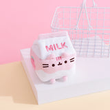 Against a pink background and yellow surface, Pusheen Strawberry Milk Sips Plush is seated on a white pedestal with a small white wire basket in the background. The plush is in the shape of a milk carton, with the word ‘Milk’ embroidered in pink stitching on the front lid of the plush, and a light pink and white wave throughout the body. Pusheen’s face is embroidered in black on the plush, with plush ears at the rim of the carton lid and pink plush feet at the bottom. 