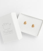 The Gold Pusheen Charm Stud earrings in a rectangular white jewelry box, the lid resting on top of the bottom left corner. The gold earrings are the only bit of color in the whole piece, and the earrings reflect the same way the silver earrings do.