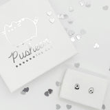 The Silver Pushen Charm Stud earrings in a jewelry box in the bottom right, with a large square jewelry box lid above them in the upper left, surrounded by silver mini hearts. All of the silver present, from the earrings, to the logo print on the lid, to the hearts, are all reflecting off light.