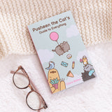 Pusheen the Cat's Guide to Everything book resting at an angle on top of a cream and white background. The book sits next to a pair of reading glasses.