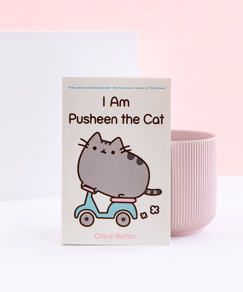 A paperback book resting against a ribbed pink pot, in front of a white and light pink background. The book cover is light yellow and features Pusheen riding a mint and pink motor scooter, with the title I Am Pusheen the Cat above her. There is a small quote in mint above the title, and the author’s name Claire Belton is written underneath in pink.