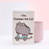 A paperback book resting against a ribbed pink pot, in front of a white and light pink background. The book cover is light yellow and features Pusheen riding a mint and pink motor scooter, with the title I Am Pusheen the Cat above her. There is a small quote in mint above the title, and the author’s name Claire Belton is written underneath in pink.