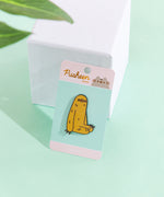 The Sloth pin in it’s packaging, leaning against a white pedestal on top a green floor, a plant leaf partially visible in the top left corner. The packaging is a round rectangular cardboard backing, a mint square in the middle and pink banners on the top and bottom. The top banner features the Pusheen Shop logo, and the bottom banner says ‘Sloth Pin’ in white.